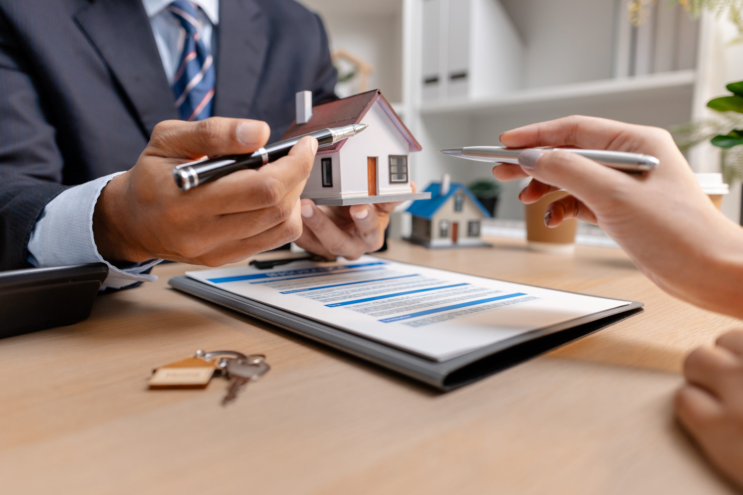 A real estate agent and client discussing property sale details at the office table, reviewing and signing home purchase contracts with miniature house models and money on the desk.