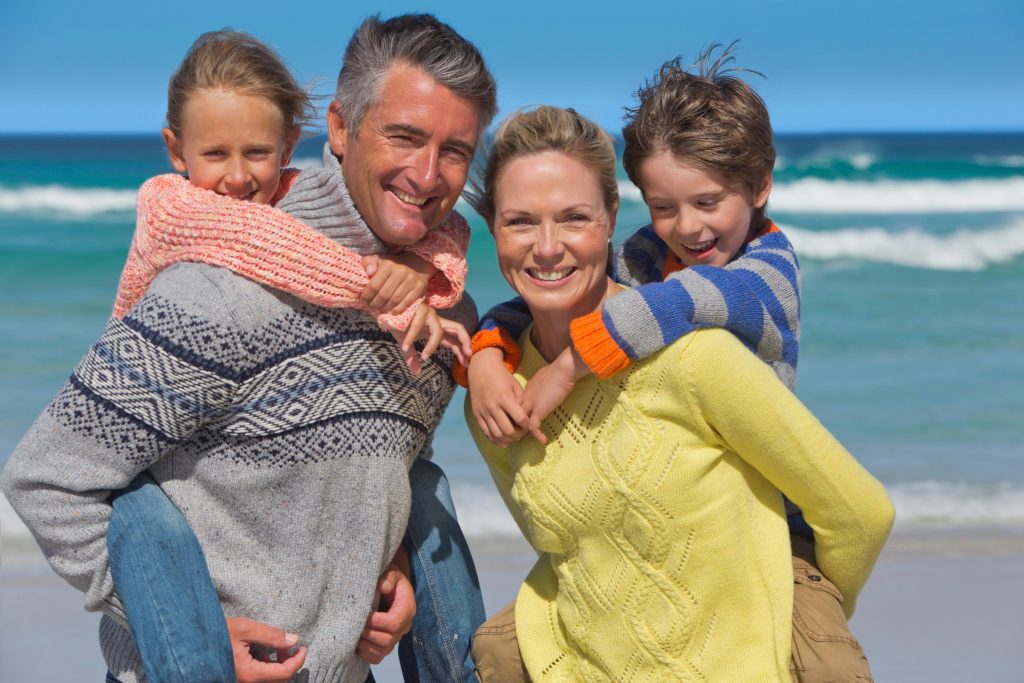 Portrait Shot Of Couple Piggybacking Their Children On A Sunny Beach.
