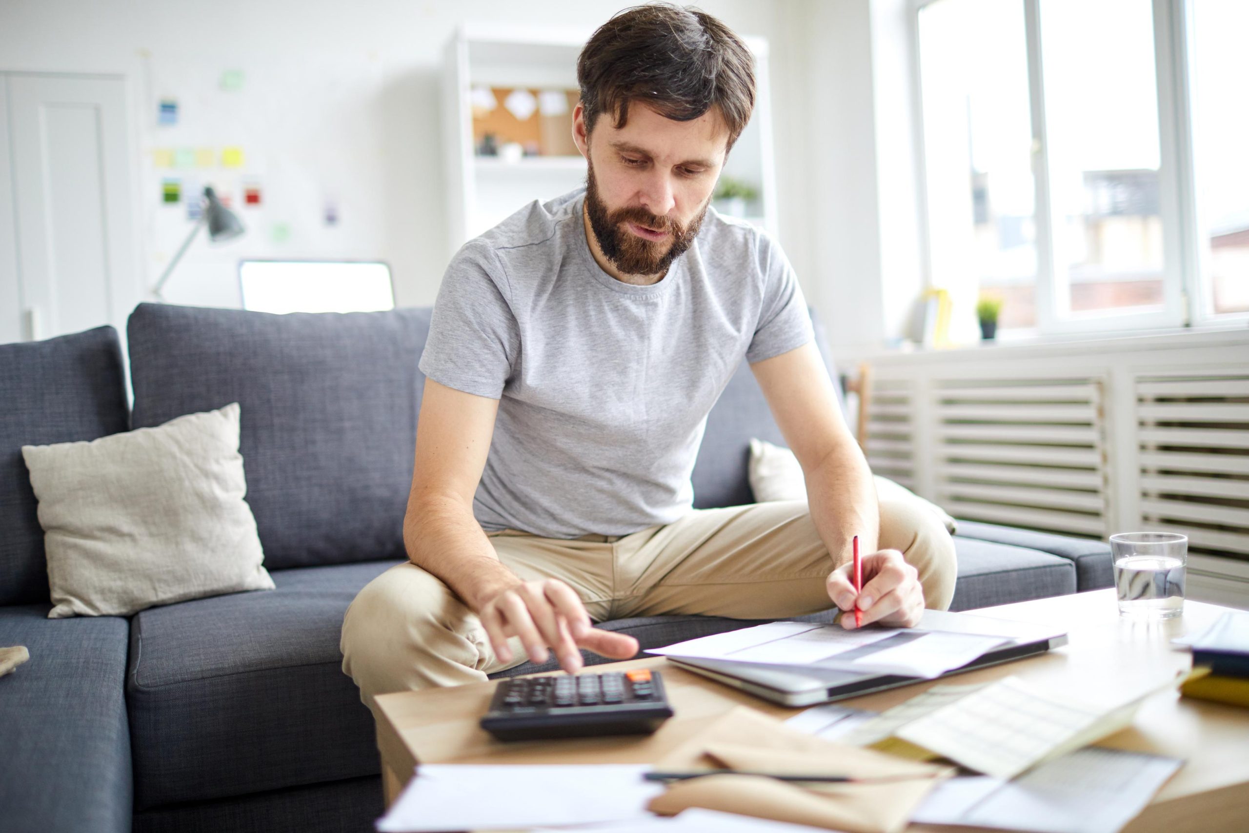 Young man in casualwear sitting on sofa in living-room and counting total sum of all payment bills