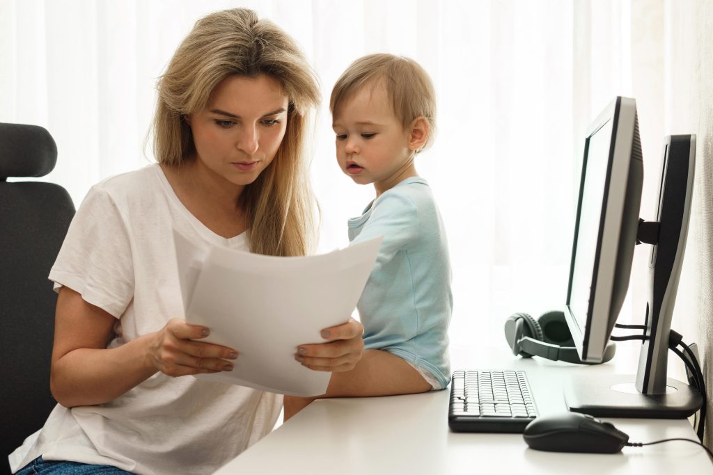 Young freelancer mother is reading some papers at her home office workplace with her curious little son sitting on the desk.