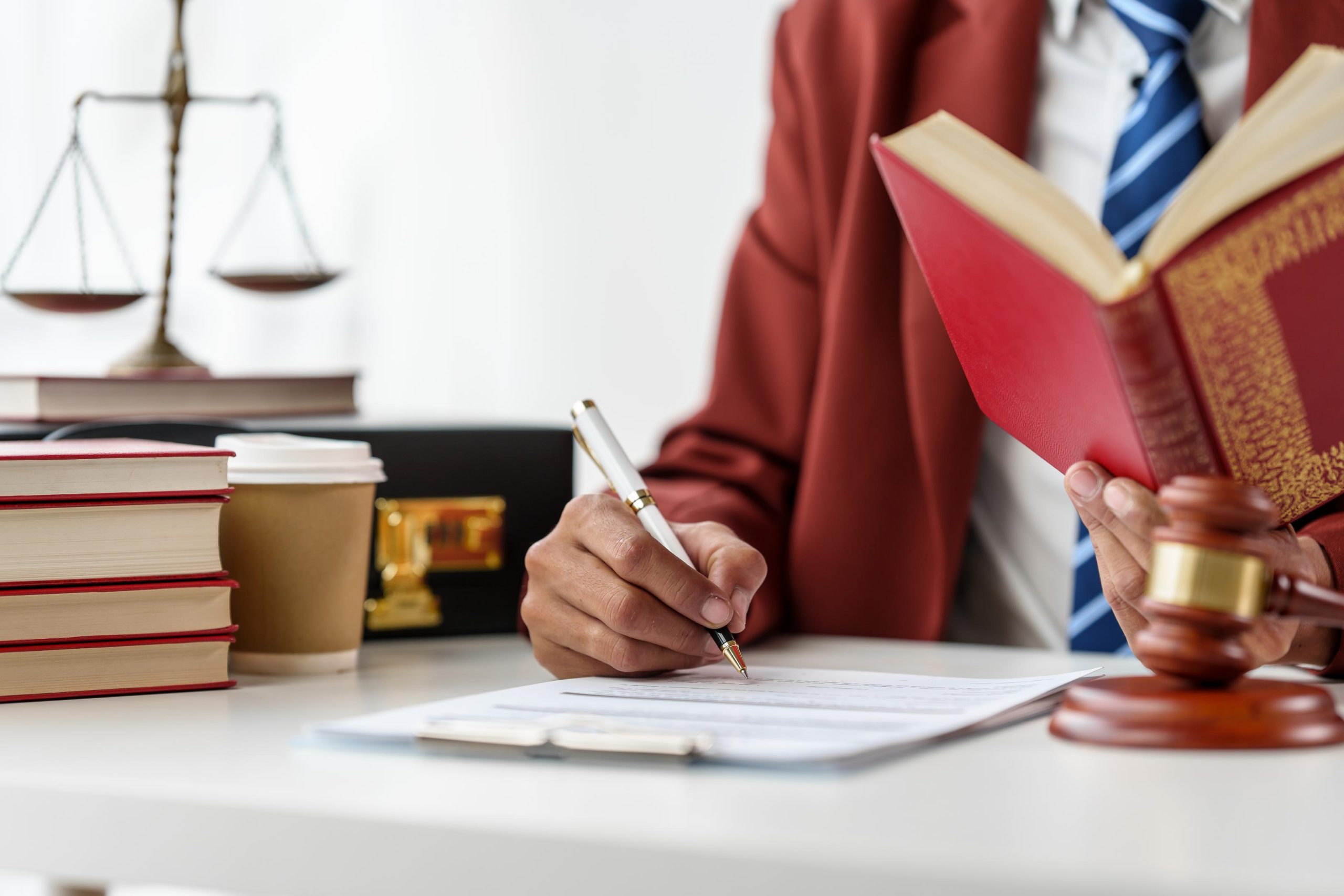 A professional sits at a white desk, signing paperwork while holding a red book.